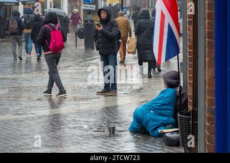 Windsor, Royaume-Uni. 14 décembre 2023. Reece, un homme qui a été rendu sans abri à la suite de la mort de son père, est assis sur le sol froid sous la pluie devant un magasin dans le riche Windsor, Berkshire, espérant que des passants lui donnent des pièces de monnaie. Polly Neate, directeur général de Shelter, a déclaré à Sky News que «le sans-abri n'est sur la liste de Noël de personne, mais 309 000 personnes passeront cette période de l'année dans une minuscule chambre d'auberge ou geler dans une porte». Crédit : Maureen McLean/Alamy Live News Banque D'Images