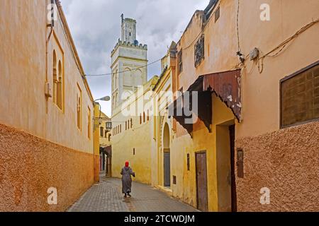 Minaret et femme musulmane portant djellaba / jillaba et hijab marchant dans l'allée de la médina jaune de la ville Meknès, Fès-Meknès, Maroc Banque D'Images