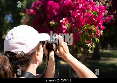 Gros plan d'une jeune femme de derrière portant une casquette rose prenant une photo avec un appareil photo reflex de fleurs violettes sur un arbre par une journée ensoleillée. Loisirs Banque D'Images