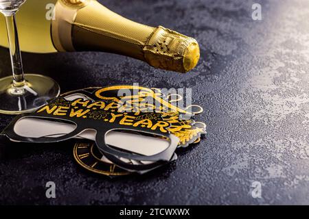 Joyeux fond de nouvelle année. Verres en papier drôles noirs et bouteille de champagne sur la table noire. Banque D'Images