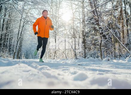 Homme de coureur de Trail souriant d'âge moyen vêtu d'une veste coupe-vent orange vif endurance course rapide forêt enneigée pittoresque pendant une journée ensoleillée gelée. S Banque D'Images