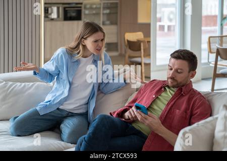 Querelle familiale. Homme concentré traîner sur le smartphone ignorant la femme insatisfaite assise sur le canapé à la maison Banque D'Images