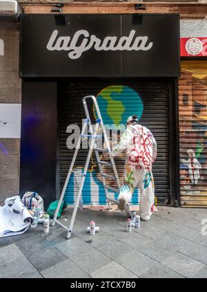 Un artiste de rue dans une combinaison peinte créant une murale colorée sur un obturateur de ville à Séville, en Espagne. Banque D'Images