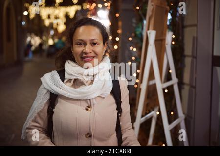 Belle femme multi ethnique regarde la caméra, sourit largement, exprimant des émotions positives, profitant de la promenade dans la belle rue décorée avec Chris Banque D'Images