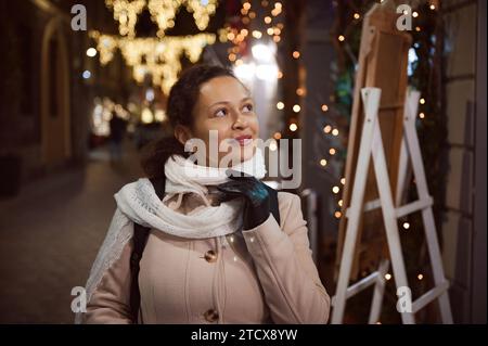 Authentique portrait de style de vie d'une charmante jeune femme en manteau beige chaud, sourires regardant de côté, admirant les décorations de Noël sur la rue en t Banque D'Images