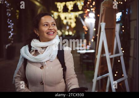 Gros plan authentique portrait de jovial jovial femme multi ethnique, en manteau chaud beige, sourit un beau sourire toothy, flânant le St élégant lumineux Banque D'Images