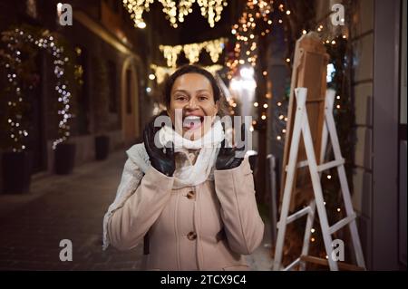 Femme multi ethnique émotive sourit regardant la caméra, profitant de l'atmosphère de Noël en plein air dans le parc des expositions la nuit, debout sur la rue de la ville Banque D'Images