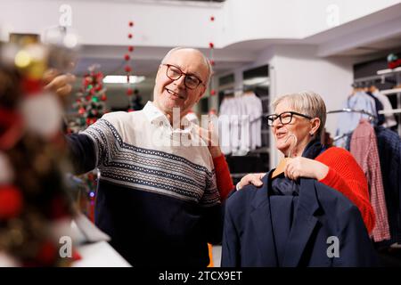 Couple senior recherchant des vêtements sur videocall dans le magasin de détail, essayant d'acheter la mesure parfaite d'article de mode pour la fête de noël. Deux personnes âgées magasinant des cadeaux de Noël pendant la saison des fêtes. Banque D'Images