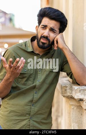 Triste homme indien malheureux pense aux préoccupations de la vie, souffre d'une situation injuste. Problème, se sentir mal, agacé, chagrin d'épuisement. Frustré déprimé arabe solitaire se tenant dans la rue de la ville. Vertical Banque D'Images