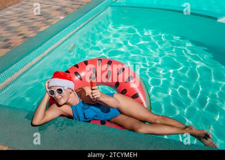 Chapeau de père Noël piscine femme. Une femme heureuse dans un bikini bleu, un chapeau de Père Noël rouge et blanc et des lunettes de soleil pose près de la piscine avec une coupe de champagne debout Banque D'Images