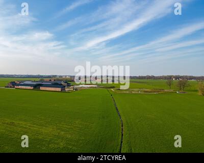 Cette image présente une perspective aérienne à couper le souffle de vastes terres agricoles sous un ciel bleu vibrant avec des nuages rusés. Les formes géométriques contrastées des bâtiments de la ferme contre les champs verts uniformes créent un motif visuellement frappant. Un chemin étroit et sinueux ou une voie navigable traverse les champs, ajoutant un élément dynamique à la composition. La scène est baignée de lumière du soleil, soulignant la vivacité des champs verts, signalant un environnement agricole prospère. Vue aérienne étendue des bâtiments agricoles au milieu de terres agricoles luxuriantes. Photo de haute qualité Banque D'Images