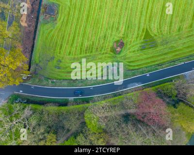 Cette photographie aérienne capture une vue panoramique d'une route asphaltée incurvée qui tranche à travers un champ verdoyant. La route est bordée par un riche feuillage, y compris un groupe d'arbres arborant des teintes automnales. Deux véhicules peuvent être vus voyageant sur la route, fournissant une sensation d'échelle et de mouvement. Le vert vif du champ est marqué par des motifs issus de l’activité agricole, contrastant avec le gris de l’asphalte. L’interaction des éléments naturels et artificiels offre un récit d’harmonie et de coexistence. Vue aérienne d'une route courbe à travers une campagne animée. Photo de haute qualité Banque D'Images