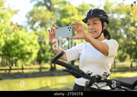 Heureuse femme d'âge moyen dans le casque de vélo s'arrête à prendre la photo du paysage avec le téléphone portable. Banque D'Images