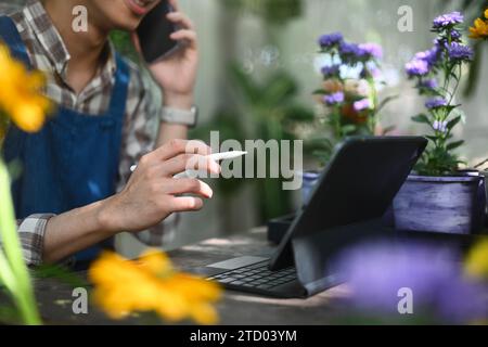 Fleuriste masculin heureux parlant sur le téléphone mobile et en utilisant un ordinateur portable pour prendre des commandes au magasin de fleurs. Banque D'Images