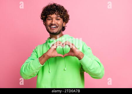 Portrait de jeune beau gars indien aux cheveux ondulés portant un sweat-shirt vert montrent le symbole du coeur à la poitrine isolé sur fond de couleur rose Banque D'Images