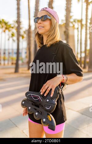 Souriant jeune femme blonde à la mode portant des lunettes de soleil et casquette debout avec des patins à roulettes en ligne dans le parc pendant le week-end Banque D'Images