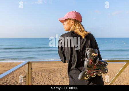 Vue arrière de la jeune femme caucasienne habillée en casuals avec des patins à roulettes alignées s'appuyant sur la rampe de cabane de sauveteur à la plage sous le ciel bleu Banque D'Images