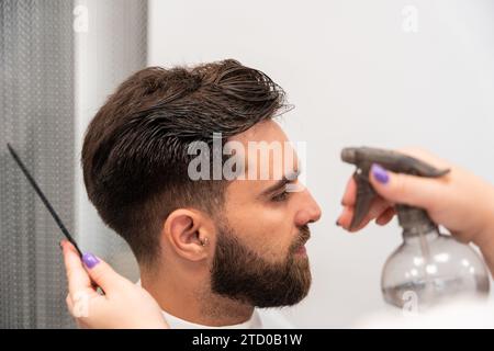 Coupez les mains de coiffeur féminin méconnaissable pulvérisant de l'eau sur les cheveux du client masculin dans le salon Banque D'Images