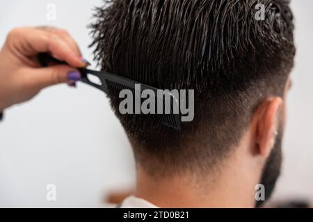 Coupe main de coiffeur féminin tenant le peigne dans les cheveux du client masculin au salon Banque D'Images