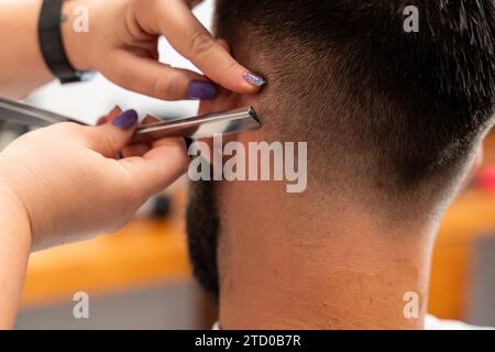 Mains recadrées méconnaissables de coiffeur féminin coiffant les cheveux du client masculin avec rasoir droit dans le salon Banque D'Images