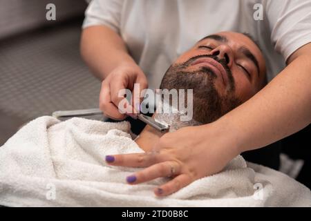 Vue à angle élevé des mains du coiffeur féminin rasant la barbe du client masculin avec rasoir droit au salon Banque D'Images