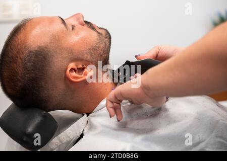 Vue latérale des mains d'un barbier coupé et méconnaissable rasant la barbe d'un client à l'œil fermé avec une tondeuse à gazon au salon Banque D'Images