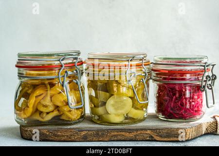 Trois bocaux en verre remplis de chou fermenté, de poivrons épicés et de concombres sur une planche de bois rustique sur fond gris. Banque D'Images