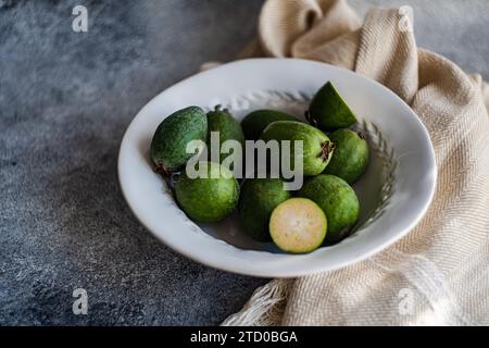 Fruits de feijoa mûrs disposés soigneusement dans un bol en céramique blanche, avec un fond de tissu texturé. Banque D'Images