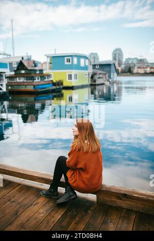 Une femme rousse contemplative est assise sur un quai en bois, contemplant les eaux sereines entourées de maisons flottantes vibrantes à Victoria, île de Vancouver, B. Banque D'Images