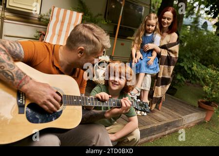 homme tatoué jouant de la guitare acoustique à gai rousse gai près de la famille et la maison de remorque moderne Banque D'Images