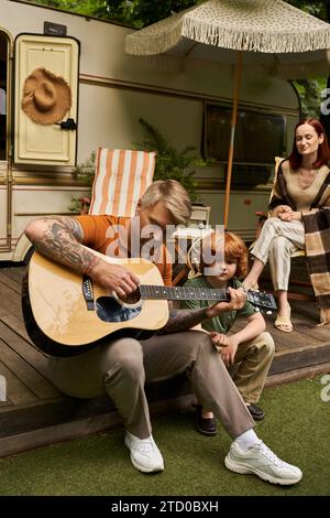 homme tatoué jouant de la guitare acoustique à son fils souriant près de la famille et la maison de remorque, moments de liaison Banque D'Images