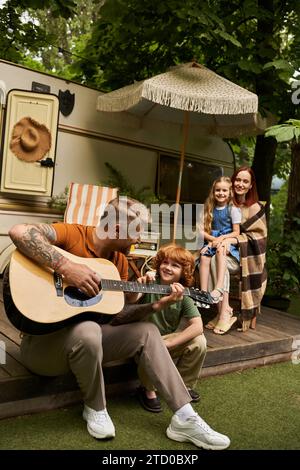 homme tatoué jouant de la guitare acoustique à fils roux heureux près de la famille et la maison de remorque moderne Banque D'Images