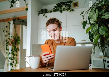 Souriant jeune femme dans des vêtements décontractés et avec des dreadlocks regardant l'écran du téléphone portable tout en étant assis à la table de cuisine avec tasse à café pour ordinateur portable et Banque D'Images