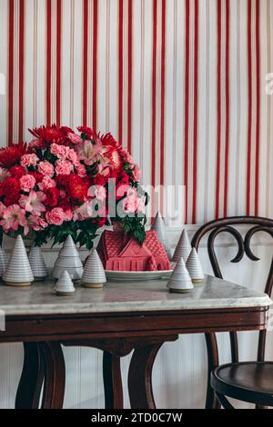 Une table en bois vintage avec un bouquet vibrant de fleurs rouges et roses, complétée par des arbres blancs décoratifs et une petite maison rose, posée contre un St Banque D'Images