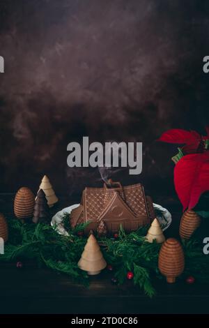 Une maison fantaisiste en pain d'épice au chocolat avec de la fumée s'élevant de la cheminée, entourée de verts de sapin festifs, de poinsettia et de bougies en cire d'abeille. Banque D'Images