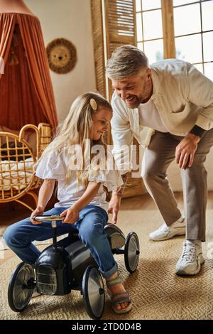 père attentionné aidant la fille heureuse chevauchant la voiture de jouet dans le salon confortable à la maison, jouant ensemble Banque D'Images