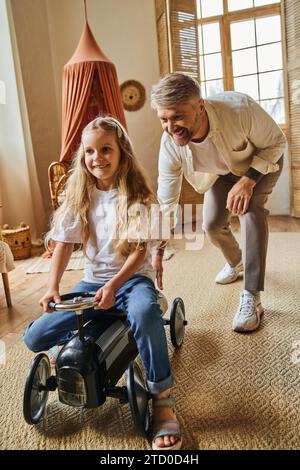 homme heureux aidant fille joyeuse chevauchant la voiture jouet dans le salon moderne à la maison, jouant ensemble Banque D'Images