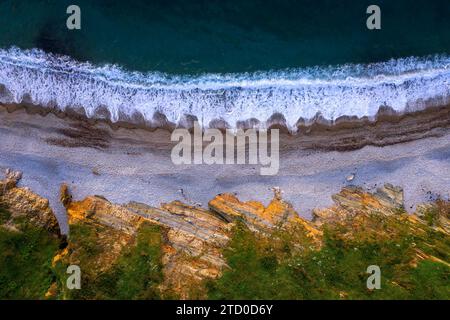 Prise de vue aérienne capturant la dynamique côte occidentale des Asturies en Espagne, avec des vagues s'écrasant sur la plage de galets et des affleurements rocheux. Banque D'Images