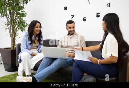 Heureux collègues masculins et féminins multiraciaux avec ordinateur portable et rapports planifiant la stratégie d'entreprise tout en étant assis sur le canapé au bureau Banque D'Images