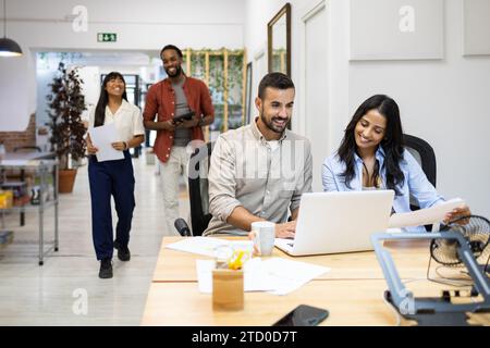 Souriant divers collègues masculins et féminins discutant du plan sur ordinateur sans fil tout en étant assis au bureau dans un bureau créatif avec des gens d'affaires à l'arrière Banque D'Images