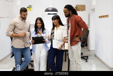 Heureux collègues masculins et féminins habillés en occasionnels discutant sur tablette numérique tout en se tenant dans la salle de conseil dans un bureau créatif Banque D'Images