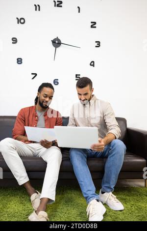 Collègues masculins multiraciaux habillés en occasionnels discutant du plan d'affaires sur le document et l'ordinateur portable tout en étant assis sur le canapé contre l'horloge sur le mur en créati Banque D'Images