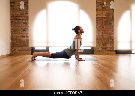 Un homme effectue un étirement de yoga sur un tapis dans une pièce avec des planchers de bois franc et des murs de briques apparentes, illuminés par la lumière naturelle. Banque D'Images