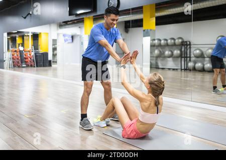 Un entraîneur personnel félicite une cliente avec un high five alors qu'elle fait des pompes abdominales dans une salle de gym, faisant preuve de travail d'équipe et de motivation. Banque D'Images