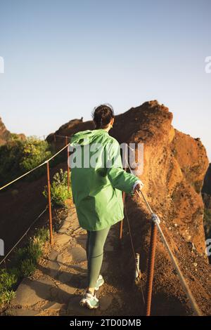 Vue arrière de la jeune femme méconnaissable habillée d'imperméable vert marchant sur le sentier par la rambarde au sommet de la montagne contre le ciel clair au lever du soleil Banque D'Images