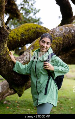 Heureuse jeune femme portant imperméable vert avec sac à dos debout près de l'arbre couvert de mousse dans la forêt pendant l'escapade de week-end Banque D'Images
