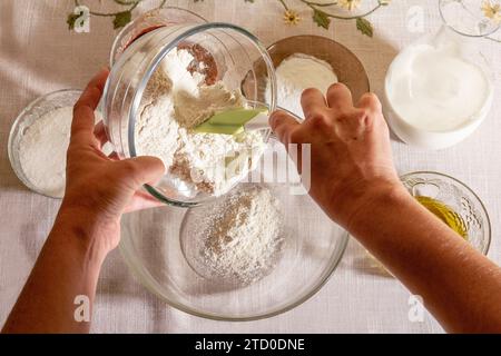 Vue de dessus des mains de la culture anonyme personne versant de la farine dans un bol en verre au milieu de divers ingrédients de cuisson sur une table avec un tableclot floral subtil Banque D'Images