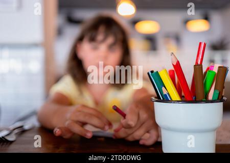 Divers crayons de couleur et stylos dans la tasse en plastique placé sur la table en bois contre fille élémentaire caucasienne passant du temps libre au restaurant Banque D'Images