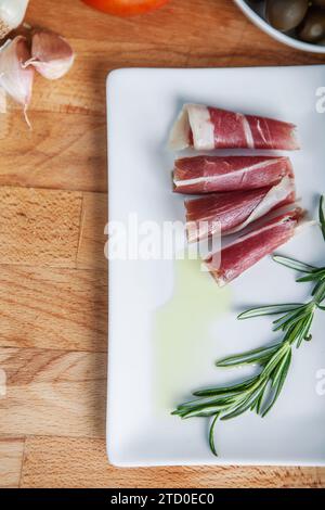 Jambon séché tranché arrosé d'huile d'olive à côté de romarin frais sur une assiette blanche, avec de l'ail et une tomate en arrière-plan. Banque D'Images