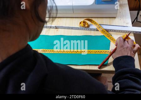 Personne préparant un morceau de tissu avec un ruban à mesurer et un crayon pour une tâche de couture à une table en bois, avec une machine à coudre en arrière-plan. Banque D'Images